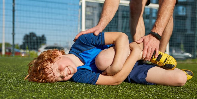 Bambino sdraiato su erba sintetica, con espressione di dolore mentre si tiene gamba