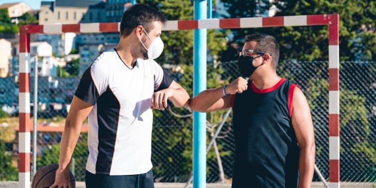 due persone con abbigliamento sportivo che indossano la mascherina mentre si salutano con il gomito su un campo da basket