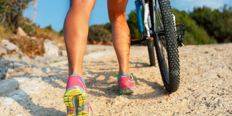 dettaglio polpacci di persona che cammina accanto a una bicicletta su un sentiero sterrato.